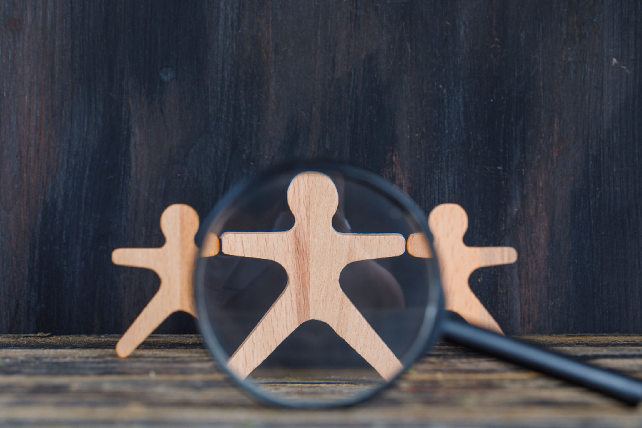 A magnifying glass held by wooden figures on a table, illustrating the focus on evaluating worker skills in hiring practices.
