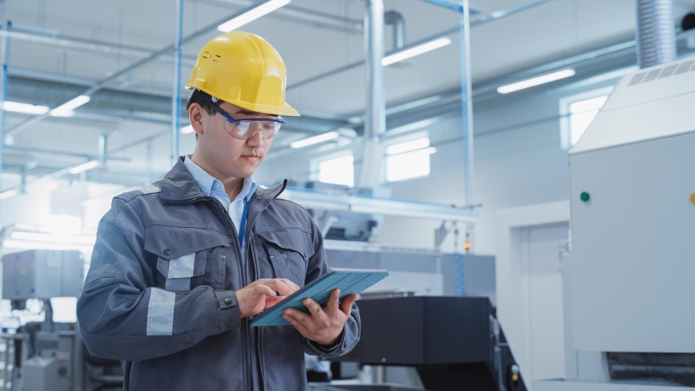 An engineer in a factory wearing a yellow hard hat and safety glasses, using a tablet to monitor operations