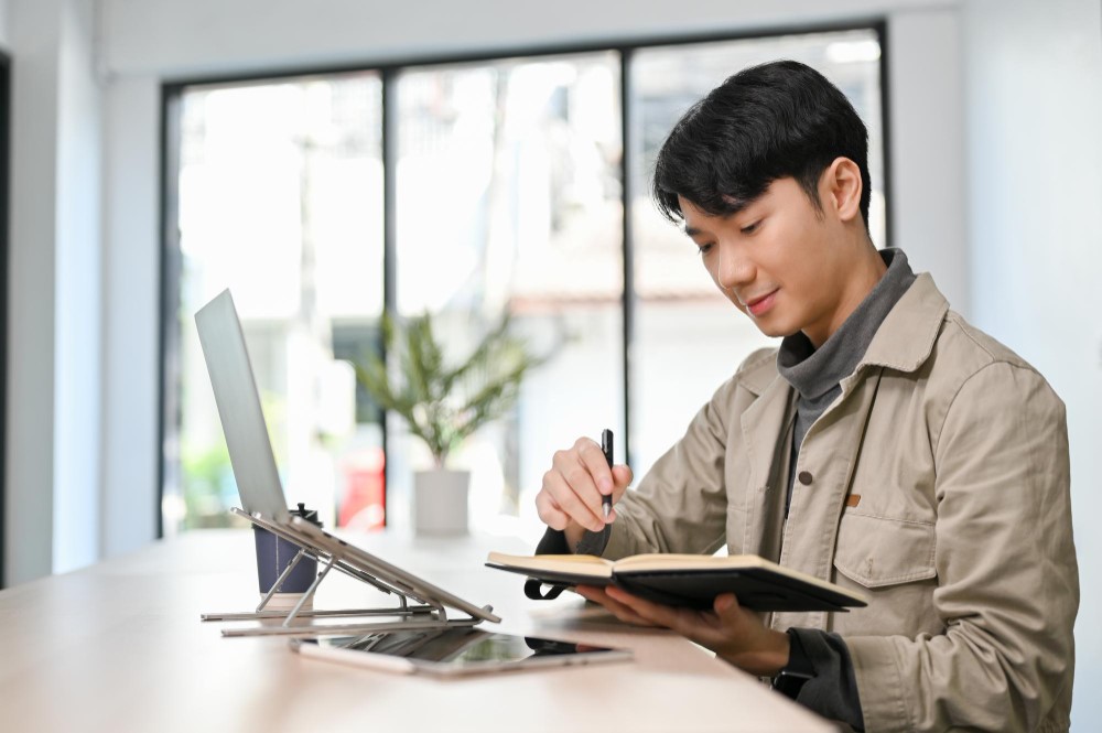 A young professional taking notes while studying with a laptop in a bright and modern workspace